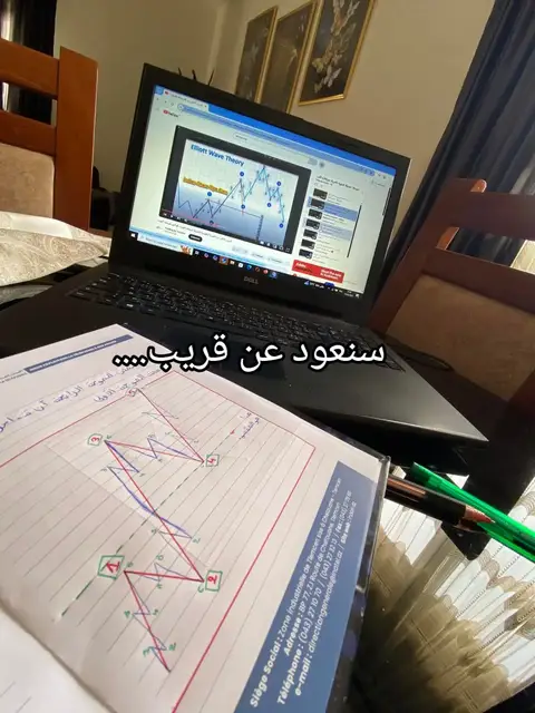A laptop displaying a chart and video related to financial trading rests on a glass table with a notebook and pen. The background includes a blurred view of a room with framed artwork.