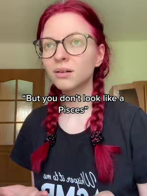 A woman with reddish hair, wearing glasses and a black shirt, poses for a video. The text overlayed on top says, "But you don't look like a Pisces".