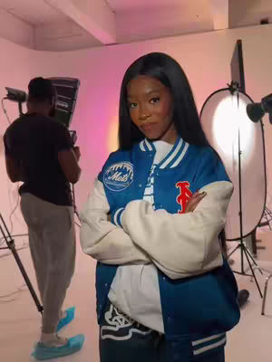 The image captures a woman with long dark hair, wearing a blue and white letterman jacket. She stands in a studio setting with lighting equipment and another person in the background. The woman has her arms crossed and appears to be posing for the camera.