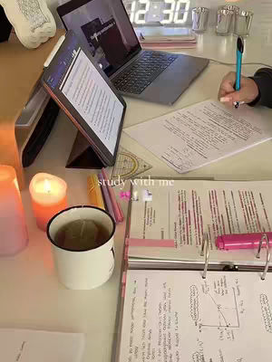 An overhead shot of a study setup featuring a laptop, tablet, and various study materials. A candle is lit to the left of the frame, and a mug with a drink sits in the foreground, along with several pens, paper sheets and open book. The colors are warm and inviting, with a focus on a well-organized workspace.