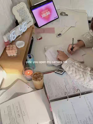 A desk covered in study materials and items, including books, papers, a tablet with a heart-shaped clock, a digital clock, candles, and a drink. In the foreground, hands are writing on a paper. The lighting is warm and inviting.