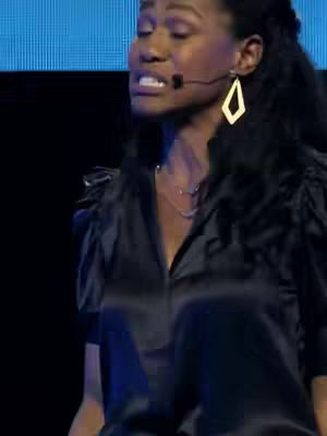 A medium shot of a woman speaking, lit by stage lighting. She has dark skin, long, curly black hair pulled back on one side, and is wearing a black top, and gold earrings. A microphone is clipped to her collar. The background is a muted blue.
