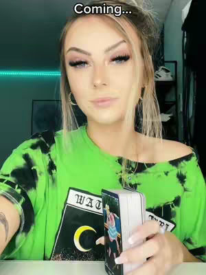 A woman with blonde hair and makeup holds a tarot card deck, looking at the camera. She is wearing a green tie-dye shirt, with a caption in the upper part of the screen.