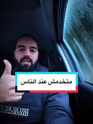 A man with a beard is giving a thumbs-up in the front seat of a car. The window is slightly blurred, and there's a text overlay in Arabic.