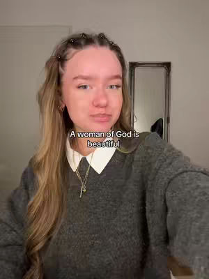 A woman with long, wavy blonde hair and a fair complexion looks directly at the camera. She's wearing a gray sweater over a white collared shirt, and a gold necklace with a cross pendant. The background is a light-colored wall and a mirror. The text overlay reads 'A woman of God is beautiful.'