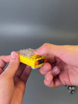 A pair of hands holding a small yellow and clear Lego taxi. The hands are in the foreground, and the background is a blurred grey.
