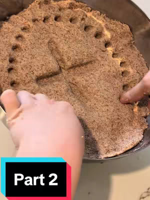 A close-up overhead shot shows a round, circular dish filled with a textured, brownish-tan substance, possibly some sort of baked dough or grain. A cross shape is clearly imprinted on the top surface, and around the edge of the substance, there are small circular indentations. Two hands are seen, one on each side of the dish, with fingers gently touching the surface. The foreground features a color-blocked bar at the bottom of the frame with the words Part 2 over a backdrop of teal, white, and black.