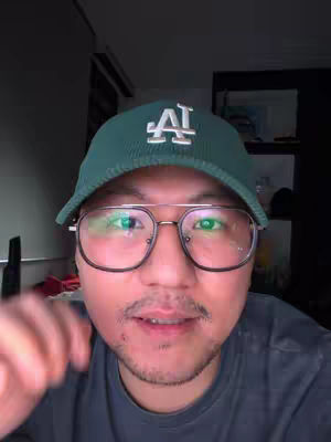 A selfie with a man wearing a green baseball cap with a white "LA" logo, a pair of glasses, and a gray t-shirt. The background is dim.