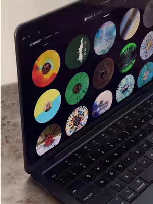 A laptop displays various colorful album art covers on its screen, resting on a surface and angled towards the viewer.