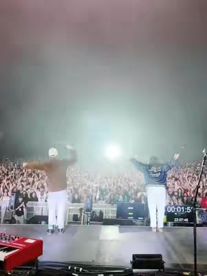 The cover image captures a nighttime scene at an outdoor event. Two figures stand on a stage in front of a vast crowd. Both figures have their arms raised in the air, with their backs towards the camera. Behind them, a large screen displays text and a timer. The background is filled with the audience, illuminated by stage lights and a bright light source behind the subjects, creating a luminous haze. The color palette is dominated by the stage, the people and the light.