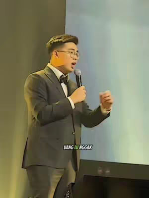 A man in a dark suit and bow tie speaks into a microphone on a stage with a bright background. Text is overlaid near the bottom of the image.