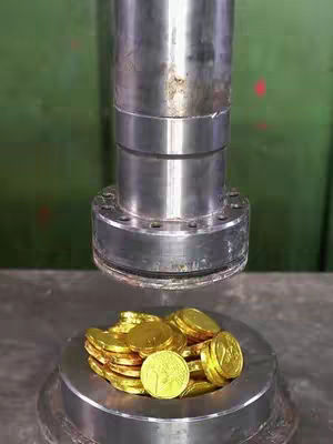 A metal press is positioned over a pile of gold coins. The press is silver, cylindrical, and has a flat, circular base. The coins are on a similar metal surface.