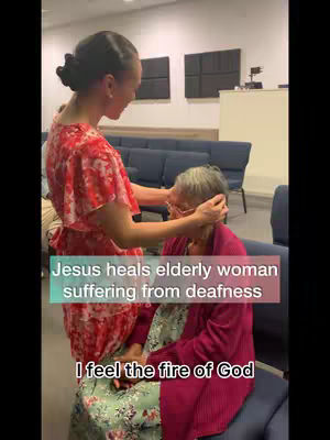 A woman in a red dress is praying over an elderly woman in a pink sweater, seated in a blue chair in front of a row of blue chairs. Text overlays indicate that Jesus healed the elderly woman of deafness.