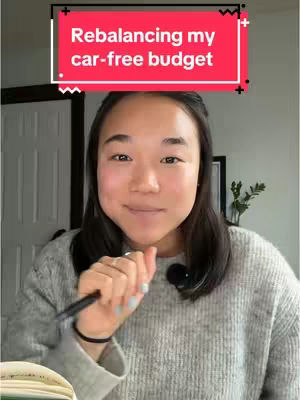 A woman with dark hair is centered in the frame, holding a pen, and smiling. Above her, a red banner with white text reads, "Rebalancing my car-free budget." The background includes a white door and a plant.