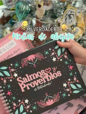 A person holds a Bible-themed journal open, with a black cover featuring pink floral designs and text. The background includes an out-of-focus Christmas tree with ornaments.