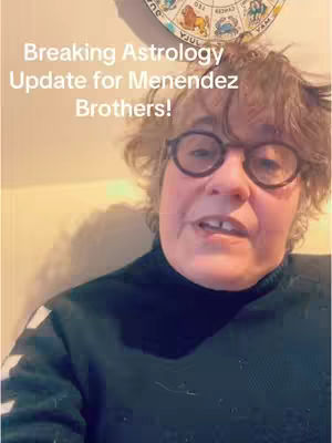A selfie-style video featuring a woman with glasses, a dark turtleneck, and short hair. Above her, a circular zodiac chart is partially visible. The background appears to be a neutral-colored wall.
