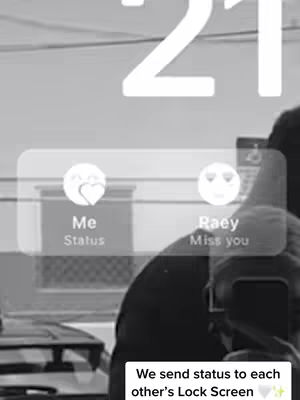 A black and white image showing the screen of a phone. The lock screen displays a date and time in a large font at the top. In the middle, there are two rounded rectangles, each containing an icon and a line of text: one with a heart icon says "Me Status", and the other with a smiley face icon that says "Raey Miss you". A person is visible in the background, holding the phone and taking the photo. The bottom of the cover image shows text, barely visible, about sending each other the statuses.