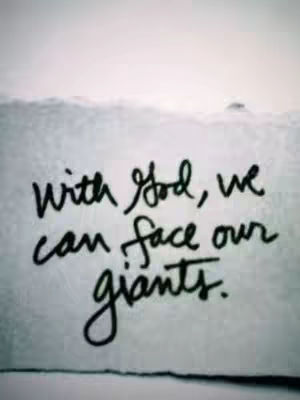 The image presents a grayscale composition. In the foreground, a slightly crumpled piece of paper bears handwritten text in dark ink. The text reads, "With God, we can face our giants." The background is a soft, blurred gradient, reminiscent of a sky or a serene setting. The overall composition is simple, conveying a sense of faith and resilience.