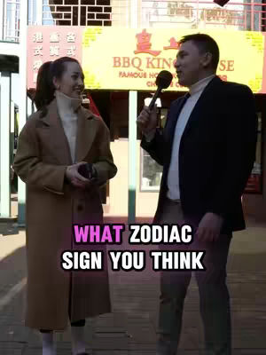 A cover image shows two people standing outside in front of stores. A woman at the left side is wearing a long brown overcoat and looking towards the man with a smile. The man at the right is holding a microphone and looking towards the woman, both smiling. In the foreground, an overlay graphic says, 'WHAT ZODIAC SIGN YOU THINK'. The background is blurred, but it appears to be a street scene with storefronts.
