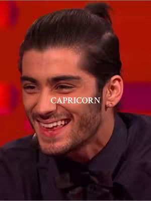 A close-up shot of a man with dark hair pulled back into a small topknot, smiling broadly. The background is a solid red and the man wears a dark shirt. White text overlayed on image has text 'CAPRICORN'