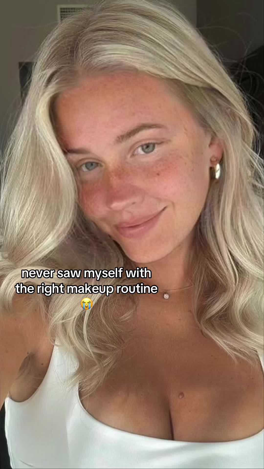 A close-up selfie of a young woman with long, wavy blonde hair. She's looking at the camera with a slight smile, skin lightly freckled, and soft lighting. The background is blurred, suggesting an indoor setting.