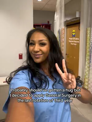 A selfie-style shot featuring a young woman in a blue medical scrub top giving a peace sign with her right hand. She has dark hair and is smiling at the camera. The background shows a hallway or corridor with doors and a sign.