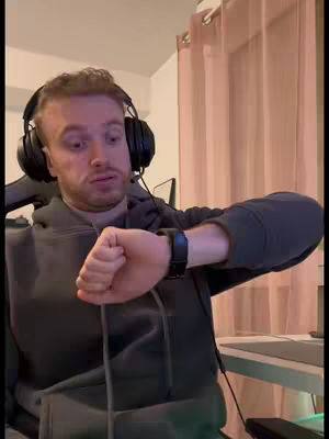 A man wearing a gray hoodie and headphones checks the time on his watch, looking concerned. The background shows a beige curtain and a corner of a room.