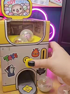 A hand holds a yellow and gray vending machine with various colorful cartoon stickers of toys, with a small, clear, plastic ball containing a figure inside.  The top has a design with a sloth.