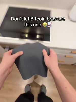 A first-person perspective shot with the creator holding a dark gray, flexible object that appears to be a mat or pad. The background contains a countertop with a large, dark-colored electronic screen or device. Text overlayed at the top indicates a focus on Bitcoin.