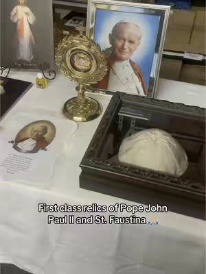 The image is a vertical shot showcasing a display of religious artifacts. The focal point is a golden reliquary, with a framed portrait of Pope John Paul II visible behind it. There are other religious items, including a photograph of a saint, and the caption 'First class relics of Pope John Paul II and St. Faustina' displayed at the bottom portion of the image.