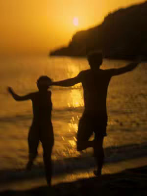 Two silhouetted figures, one male and one female, run towards the viewer on a beach. The sun is setting behind them, creating a warm, golden glow across the water. The figures have their arms outstretched in a celebratory pose. The image is slightly out of focus, enhancing the sense of movement and capturing a fleeting moment of joy.