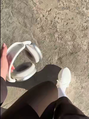 The cover image is taken from a low angle, showcasing a person's legs dressed in black leggings and white sneakers. To the left, a hand holds a pair of white headphones against a backdrop of rough, light-colored concrete or stone. The lighting is even, suggesting an outdoor setting, and the composition focuses on the person's outfit and the aesthetic of the headphones.