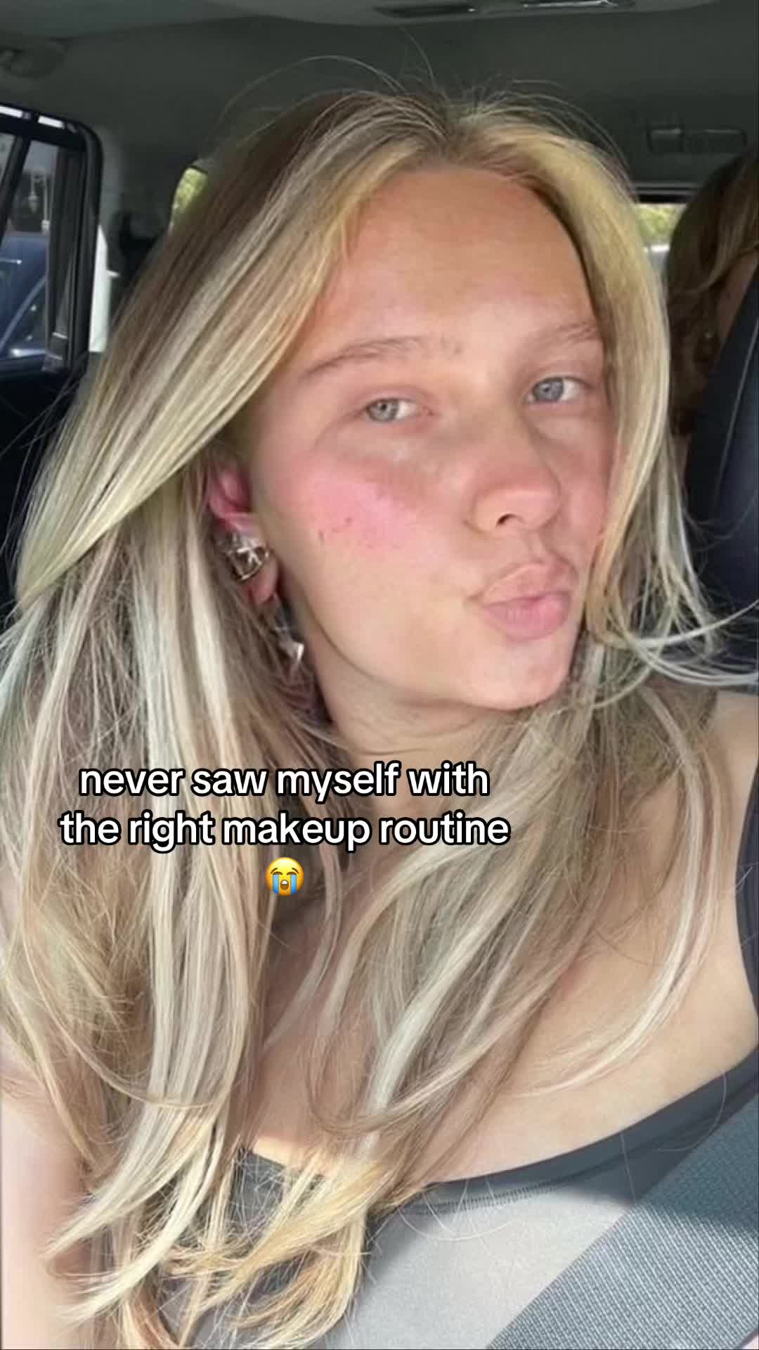 A selfie taken from inside a car features a young woman with blonde hair. She has light skin and is looking directly at the camera. The text overlay includes the phrase "never saw myself with the right makeup routine" and an emoji of a crying face.