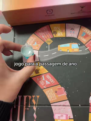 A person's hand holding a small, clear cup with a blue liquid over a board game. The board game has a circular track divided into segments, featuring game instructions and illustrations. A yellow bus graphic is prominently displayed. The color palette is warm with orange, pink, and black.