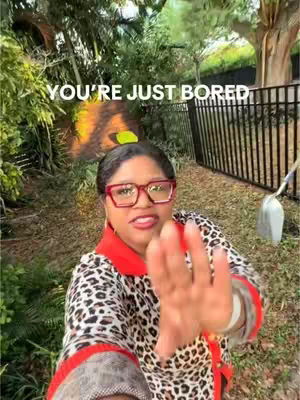 A woman with red glasses stands in a garden setting, extending her hand towards the camera. She is in a leopard print top and a red-collared shirt, with the text overlayed on the image: YOU'RE JUST BORED.