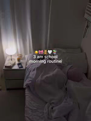 The interior of a bedroom is shown, with a bed taking up much of the frame. The bed's sheets are rumpled, suggesting someone is still in bed. A nightstand with a lamp and phone is in focus. Text overlaid on the image reads '3 am school morning routine' with related emojis.