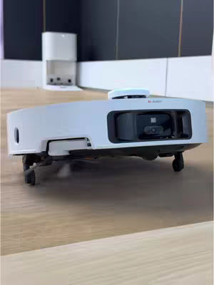 A low-angle shot of a white and black robot vacuum cleaner on a light brown wooden floor. The vacuum cleaner is rectangular with curved edges and has a prominent black sensor area in the front, and wheels are visible. In the background, a white structure resembling a charging station or base is partially visible against a dark grey wall.