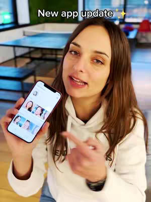 A woman is holding a phone in her left hand and is pointing at it with her right forefinger. The phone displays what appears to be a social media or app interface with several profile pictures arrayed on the screen. She is wearing a white hoodie. The background includes a table and chairs, suggesting an indoor setting The image is brightly lit, with a soft, natural light.