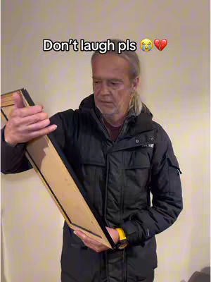 An older man with long, graying hair pulled back holds a large, light brown wooden box in front of a light beige wall. He wears a black jacket, and a yellow watch is on his wrist. Text overlays on the image.