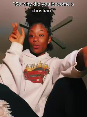 A young Black woman is centered in a medium shot, slightly angled upwards. She's wearing a white hoodie with a red and yellow graphic on it. Her dark hair is styled in a high bun. Her expression is one of slight surprise or expectation. A ceiling fan is visible in the background, and black leggings are seen in the foreground.