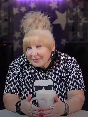 A medium shot of a woman sitting and looking at the camera. She has blonde hair in a high, messy bun and a fringe. She wears a black and white houndstooth print shirt with the Karl Lagerfeld logo on it and holds a white mug with the image of a face, possibly a stylized face. She wears several rings and bracelets. Behind her is a slightly blurred background.
