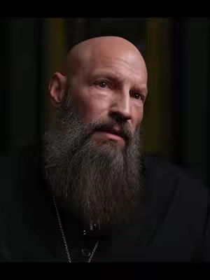 A medium shot of a bald man with a full, grey beard. He wears a dark shirt and a silver chain. The background is blurred dark.