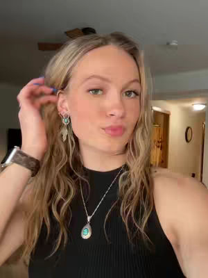 A young woman with blonde hair and fair skin is posing for a selfie. She is dressed in a black top and has long, wavy hair. She's wearing earrings and a necklace with a turquoise pendant. The background shows the inside of a home.