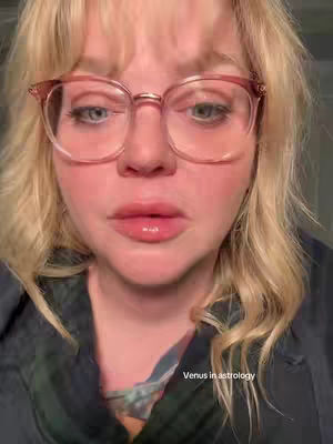 A close-up shot of a woman with blonde hair, wearing pink-framed glasses, and looking towards the camera with a neutral expression. The background is a soft, blurred environment, and overlaid at the bottom right is text that reads 'Venus in astrology'.