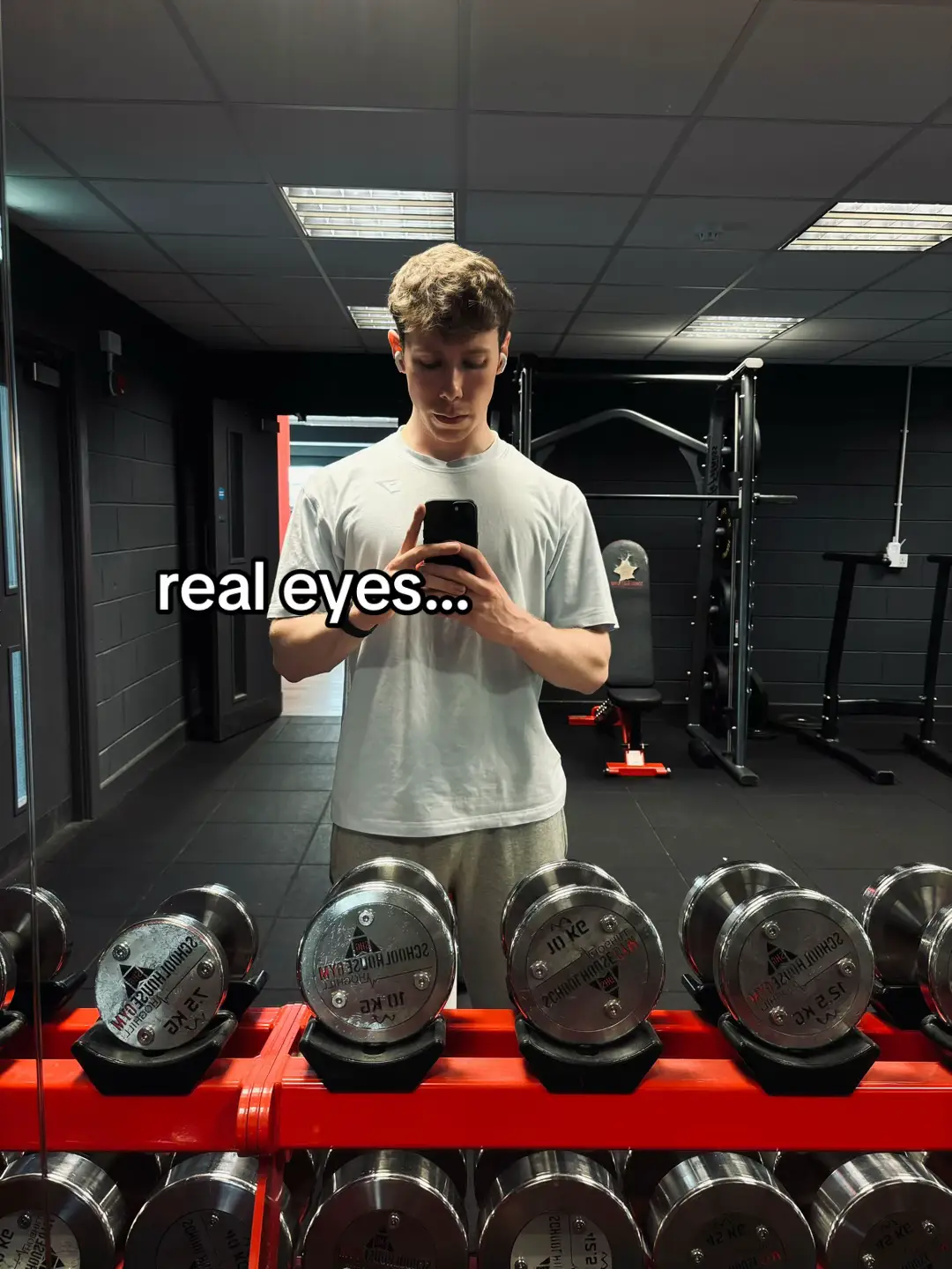 A person in a gym, standing in front of a mirror holding a phone. The person is wearing a light-colored t-shirt and gray pants. Dumbbells are visible in the foreground.