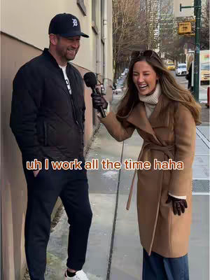 A street interview scene with a man and a woman. The man, wearing a baseball cap and black jacket, holds a microphone. The woman, in a brown coat, laughs while holding the microphone. The background shows a city street with a building and some trees.