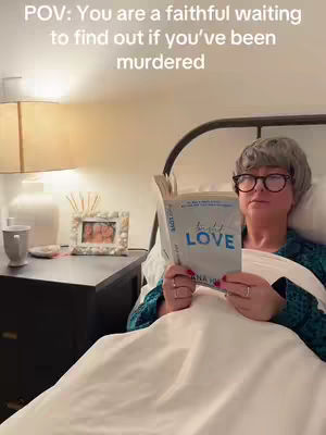 A woman reads in bed, propped up against pillows, holding a book up to the camera. She wears glasses and is under a white sheet, a bedside table with a lamp sits beside her. The overall color scheme is warm and inviting with soft lighting.