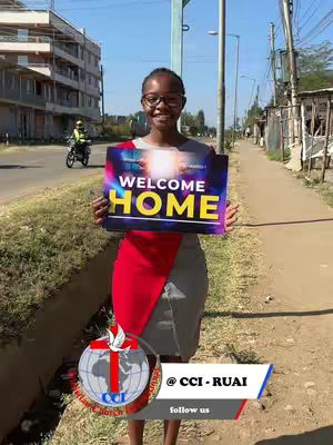 A woman wearing glasses stands on a sidewalk holding a sign that says "WELCOME HOME," with a church logo and contact information at the bottom, in front of a street with buildings and a passing motorcycle. The woman is smiling, and the overall tone is welcoming and positive.