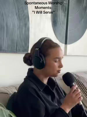 A woman with sleek dark hair, pulled back into a bun, wearing a black button-down shirt and over-ear headphones. She's holding a microphone close to her mouth, eyes closed, as she sings. Behind her, a mix of abstract art and decor is visible.