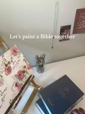A top-down shot shows a bright, well-lit workspace with a white tabletop. Several items are arranged, including an open Bible with floral artwork, a closed blue Bible, and a jar of paintbrushes. The text overlay reads "Let's paint a Bible together."